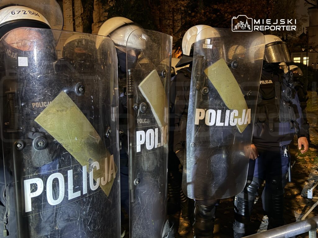 Funkcjonariusze policji w kaskach i z tarczami stoją w szeregu nocą na ulicy.