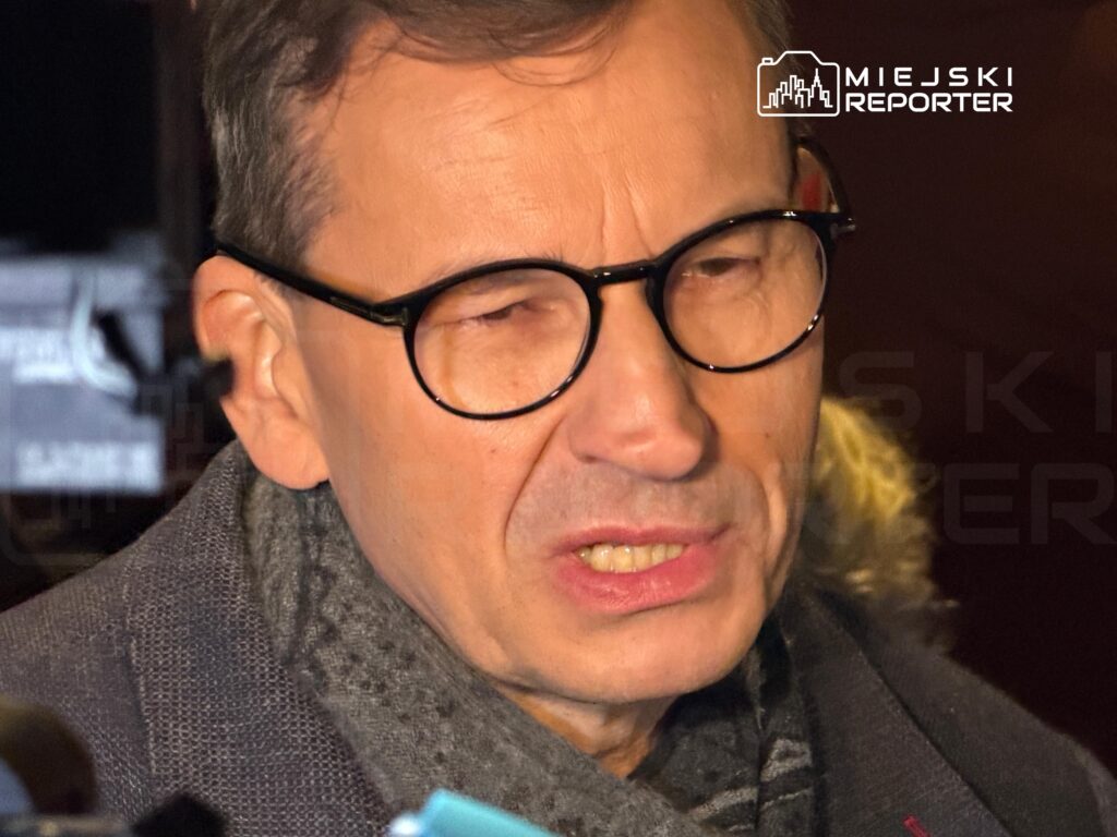 Mężczyzna w czarnych okularach i szarym szaliku podczas rozmowy z reporterami, w tle logo Miejski Reporter.