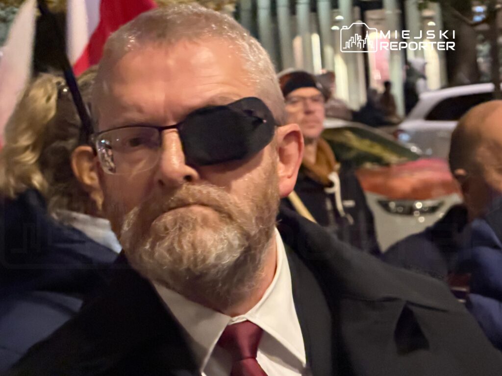 Brodaty mężczyzna w okularach z zasłoniętym jednym okiem stoi wśród ludzi na ulicy nocą.