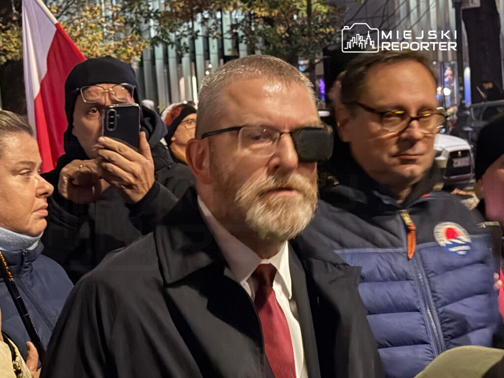 Brodaty mężczyzna w okularach i garniturze idzie w tłumie, obok osoba nagrywa go telefonem; w tle polska flaga.
