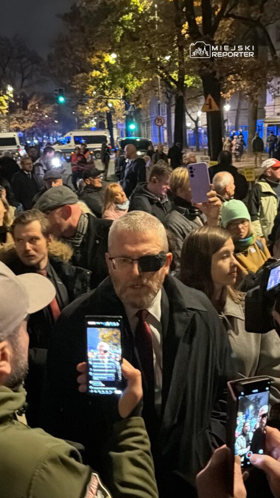 Mężczyzna w okularach przeciwsłonecznych otoczony przez tłum z telefonami na oświetlonej, zadrzewionej ulicy nocą.