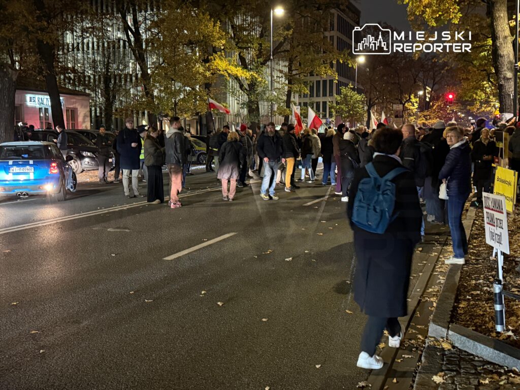 Grupa ludzi z polskimi flagami gromadzi się wieczorem na ulicy wśród samochodów i jesiennych drzew.