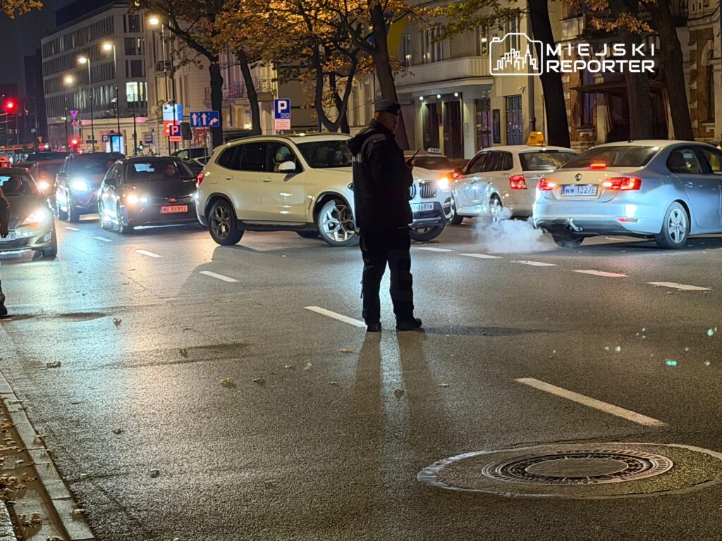 Policjant kieruje ruchem pośród samochodów na oświetlonym nocą skrzyżowaniu.