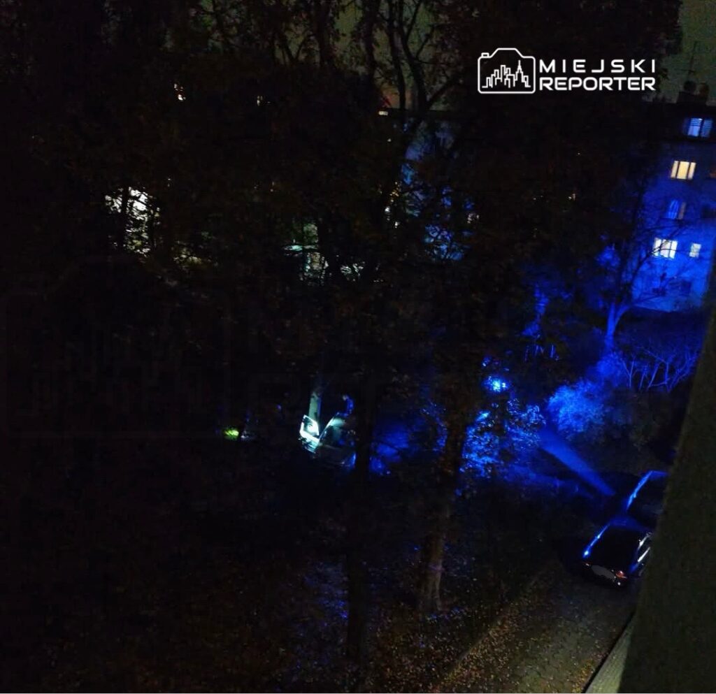 Nocna akcja służb z niebieskimi światłami przy zaparkowanych samochodach, widziana przez drzewa przy budynku mieszkalnym.