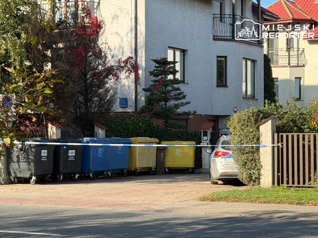 Kolorowe kontenery na odpady stoją przed budynkiem mieszkalnym, obok zaparkowanego samochodu osobowego.