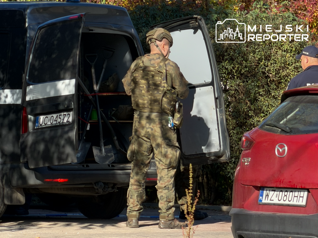 Mężczyzna w mundurze wojskowym stoi przy otwartych drzwiach dostawczego vana, obok zaparkowanego czerwonego auta.