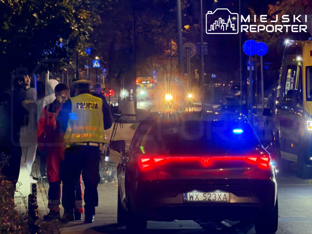 Policjant i ratownicy stoją przy radiowozie z włączonymi światłami na nocnej ulicy.