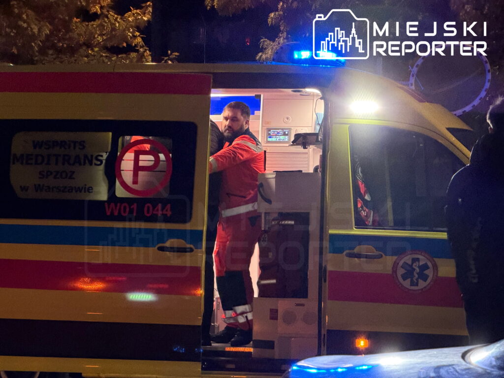 Ratownik medyczny w czerwonym uniformie stoi w otwartych drzwiach karetki z włączonymi niebieskimi światłami.