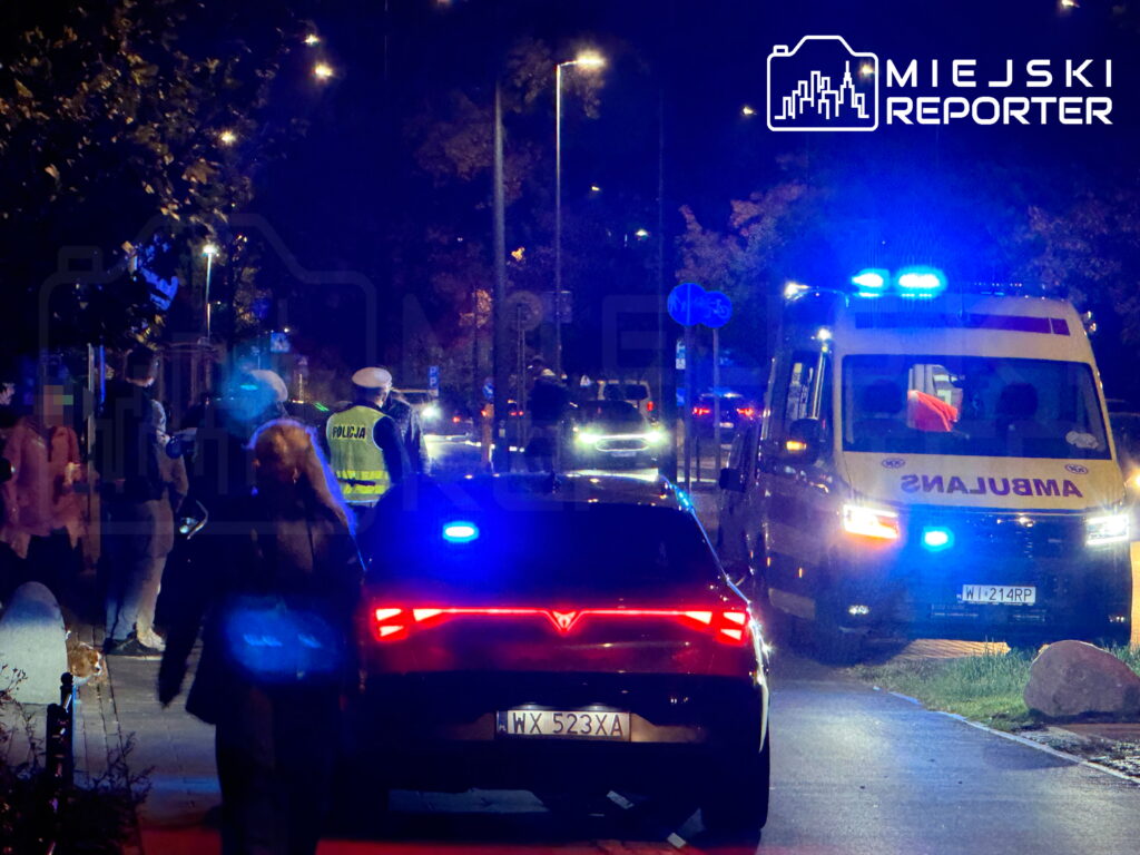 Radiowóz policyjny i karetka z włączonymi sygnałami świetlnymi na nocnej ulicy, obok policjanci i piesi.