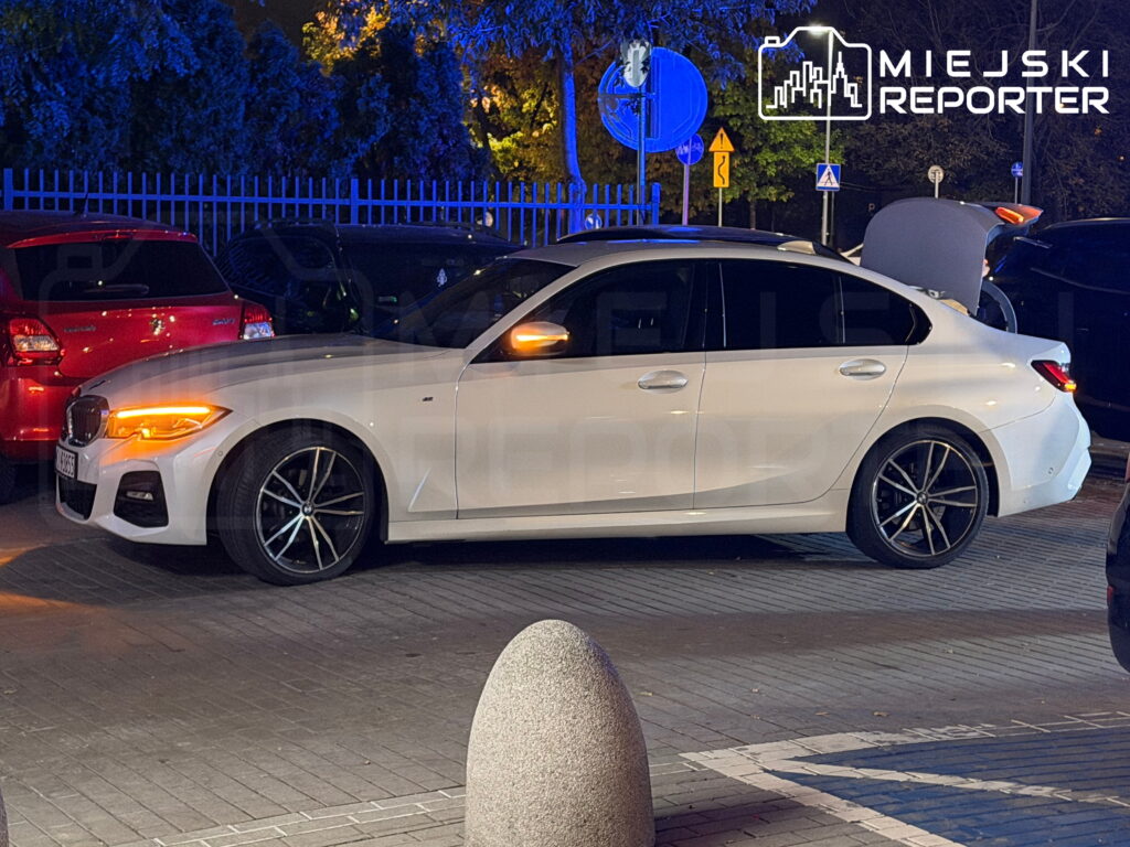 Biały sedan BMW stoi nocą na chodniku obok zaparkowanych samochodów.