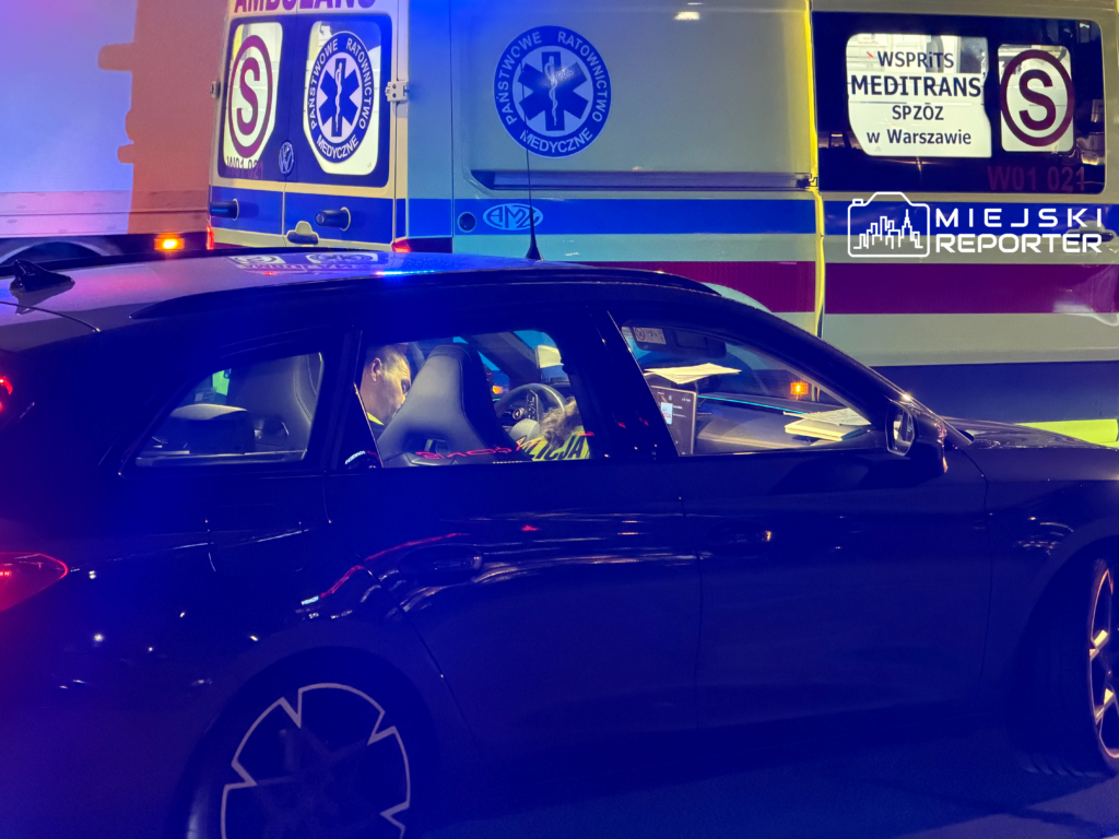 Ciemne kombi z uszkodzonym bokiem stoi przed karetką z włączonymi światłami, nocą na ulicy.