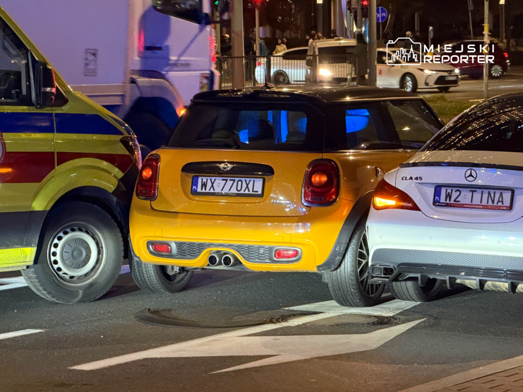 Żółty Mini i biały Mercedes stoją przy żółtym pojeździe służb na nocnej ulicy, na oznakowanym pasie jezdni.