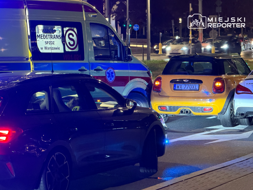 Karetka i dwa samochody stoją na skrzyżowaniu nocą, z włączonymi niebieskimi światłami pojazdu uprzywilejowanego.