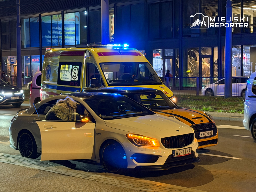 Żółty ambulans z włączonymi niebieskimi światłami stoi za białym samochodem osobowym na ulicy nocą.