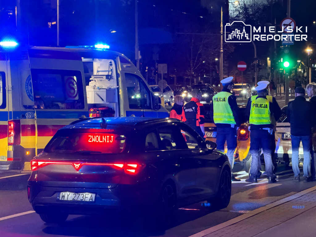 Policjanci w odblaskowych kamizelkach stoją przy karetce i samochodach na nocnym skrzyżowaniu; na aucie napis „ZWOLNIJ