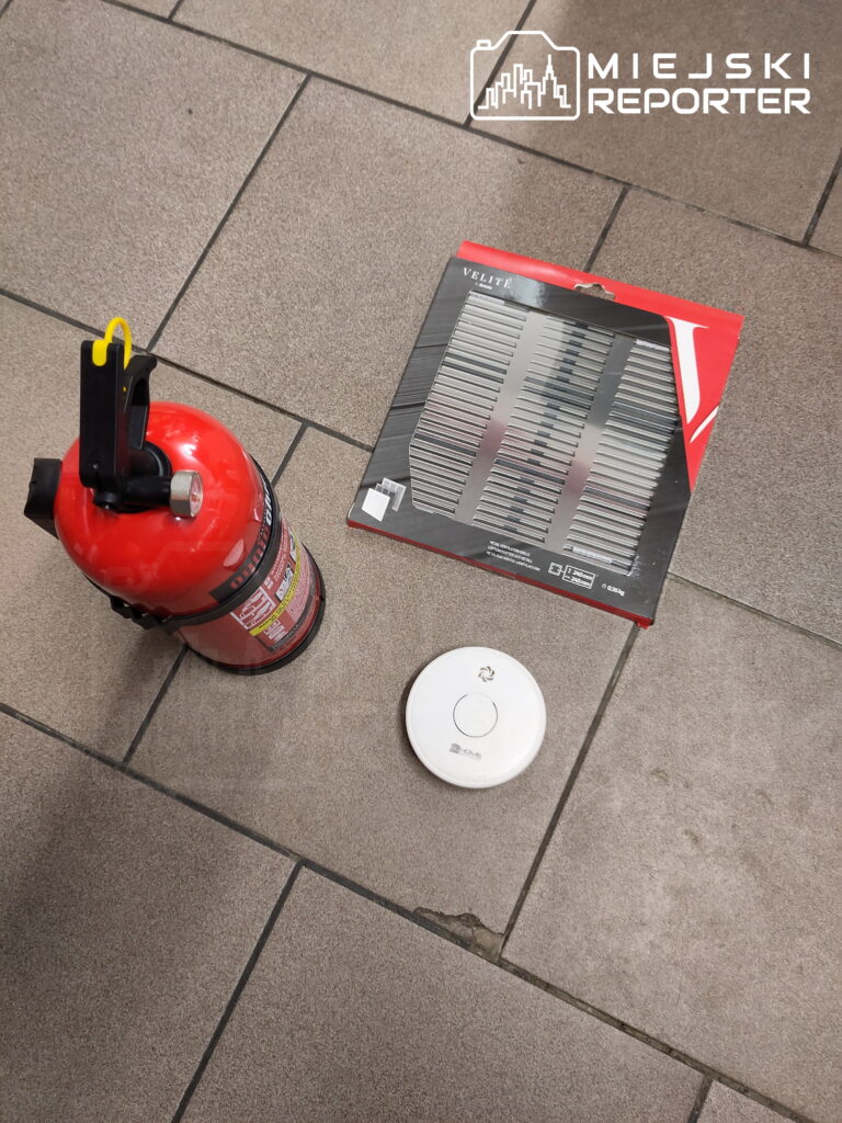 Czerwona gaśnica, opakowanie z metalową kratką i biały czujnik dymu leżą na płytkach podłogowych.