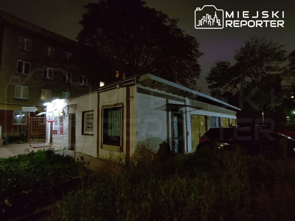 Niski, parterowy budynek z kratami w oknach i zaroślami przed wejściem, sfotografowany nocą przy bloku mieszkalnym.