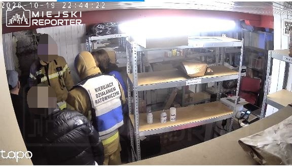 Ratownicy w odzieży ochronnej stoją w ciasnym pomieszczeniu przy metalowych regałach z pojemnikami.