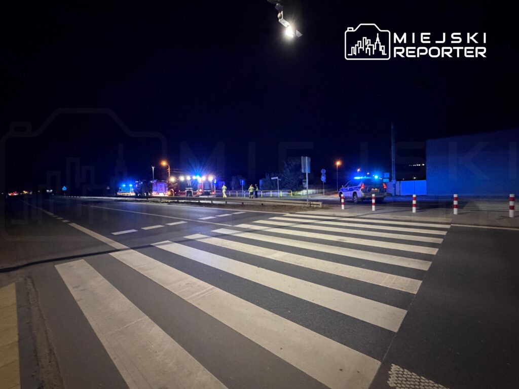 Nocne działania służb przy drodze z przejściem dla pieszych, obok pojazdów z włączonymi niebieskimi sygnałami świetlnymi.