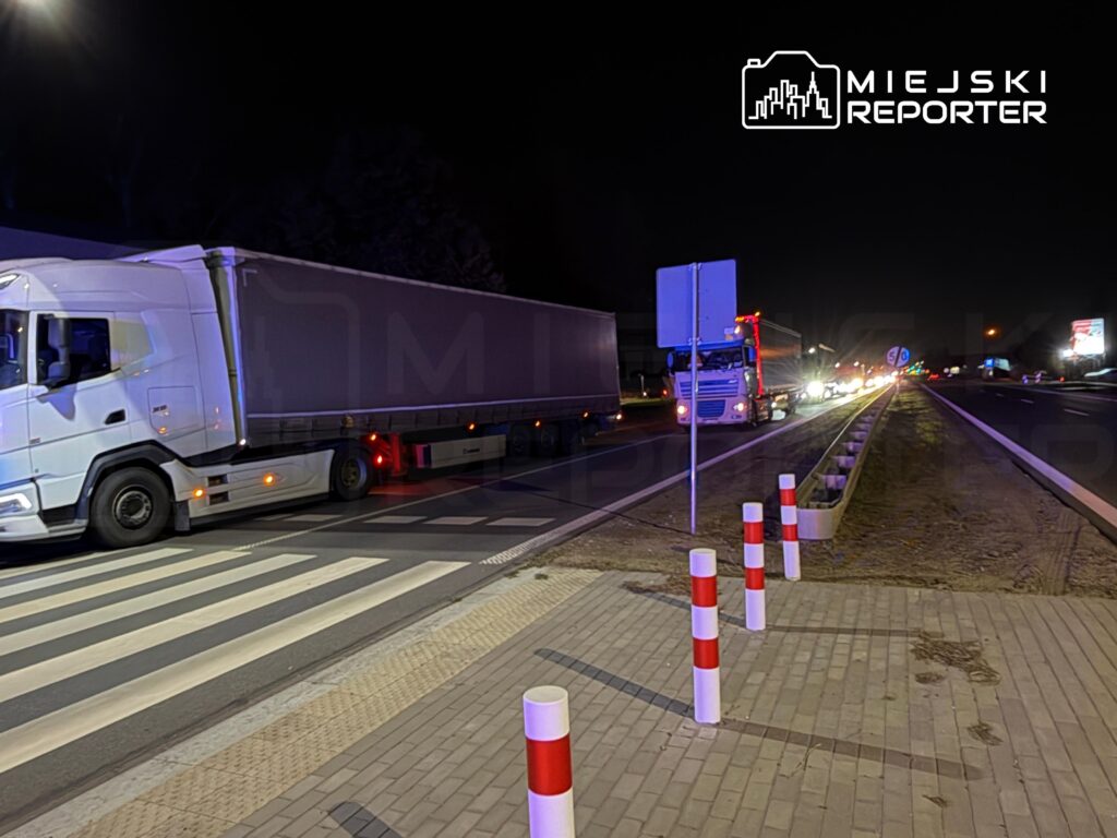 Ciężarówki stoją nocą na drodze przy przejściu dla pieszych i czerwono-białych słupkach.