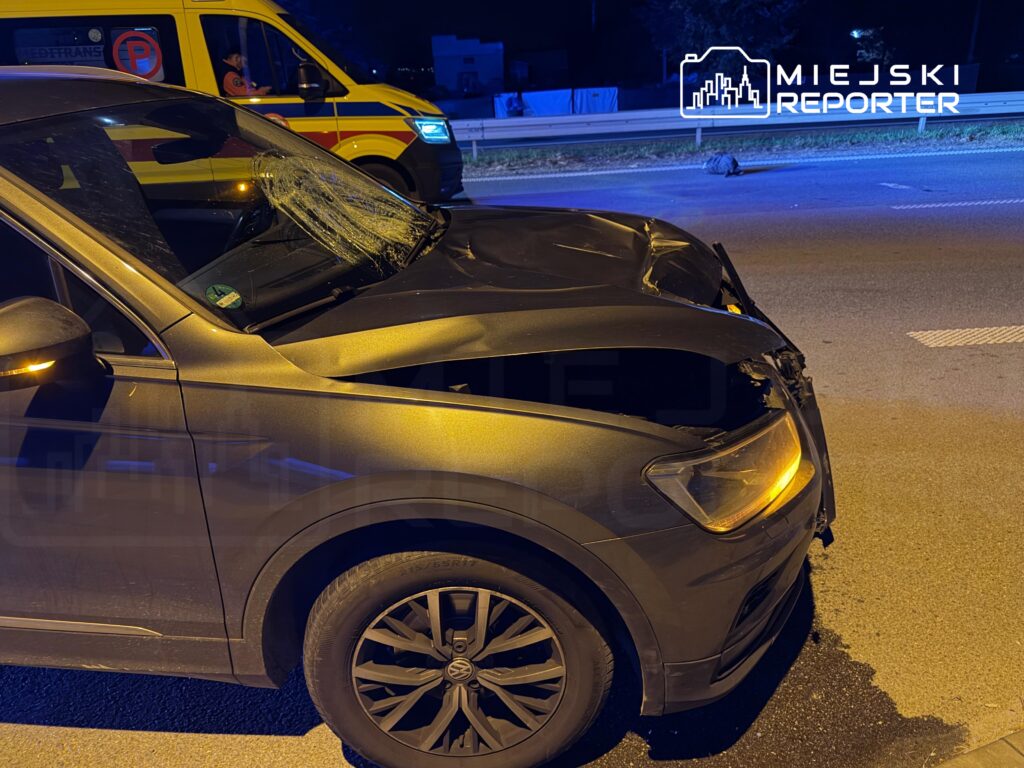 Uszkodzony samochód osobowy z rozbitą przednią szybą i zgniecioną maską na nocnej drodze, obok stoi karetka pogotowia.