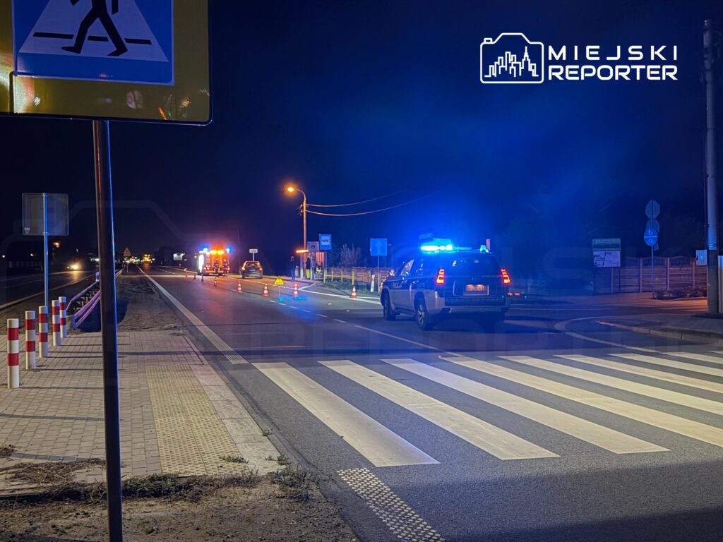Radiowóz z włączonymi niebieskimi światłami stoi na nocnej drodze przy przejściu dla pieszych, w tle pojazd służb.