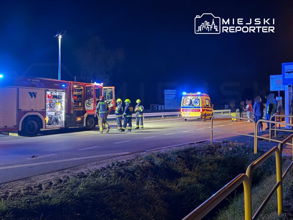 Strażacy stoją przy wozie strażackim, a karetka i służby zabezpieczają nocne zdarzenie na drodze.