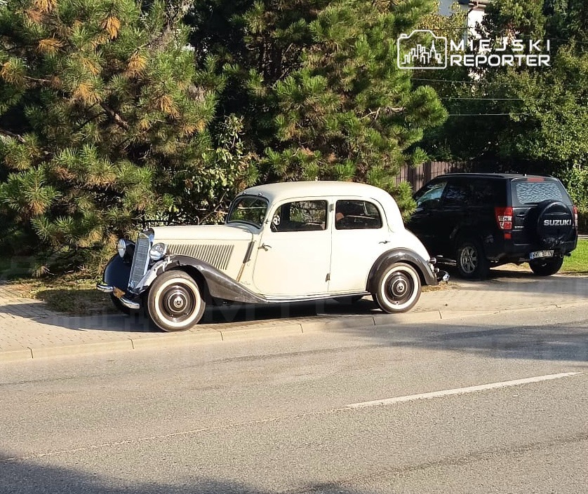 Jasny zabytkowy samochód zaparkowany przy ulicy, za nim czarny SUV i gęste drzewa.