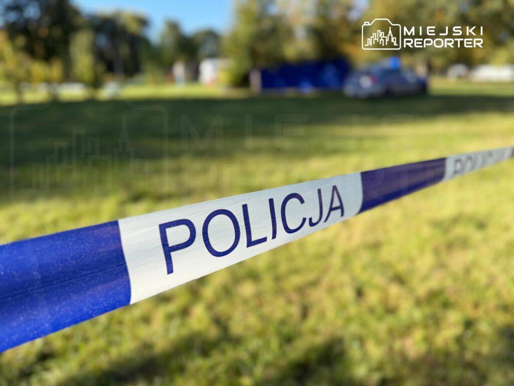 Taśma policyjna z napisem „POLICJA” odgradza trawiasty teren; w tle widać rozmyte drzewa i pojazdy.