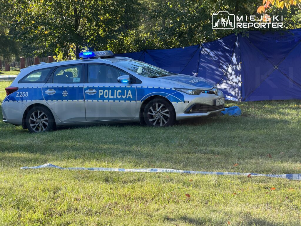 Oznakowany samochód policyjny z włączonymi niebieskimi światłami stoi na trawie przy niebieskim namiocie i taśmie policyjnej.