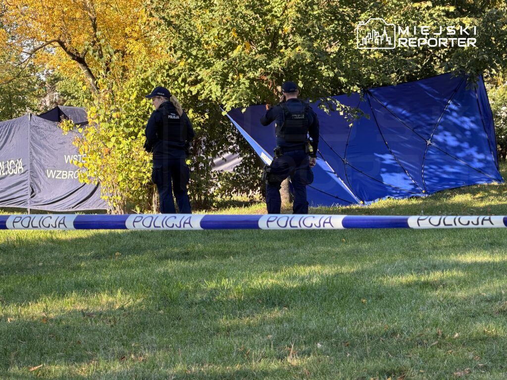 Dwóch policjantów stoi przy niebieskich namiotach za taśmą policyjną, pod drzewami na trawniku.