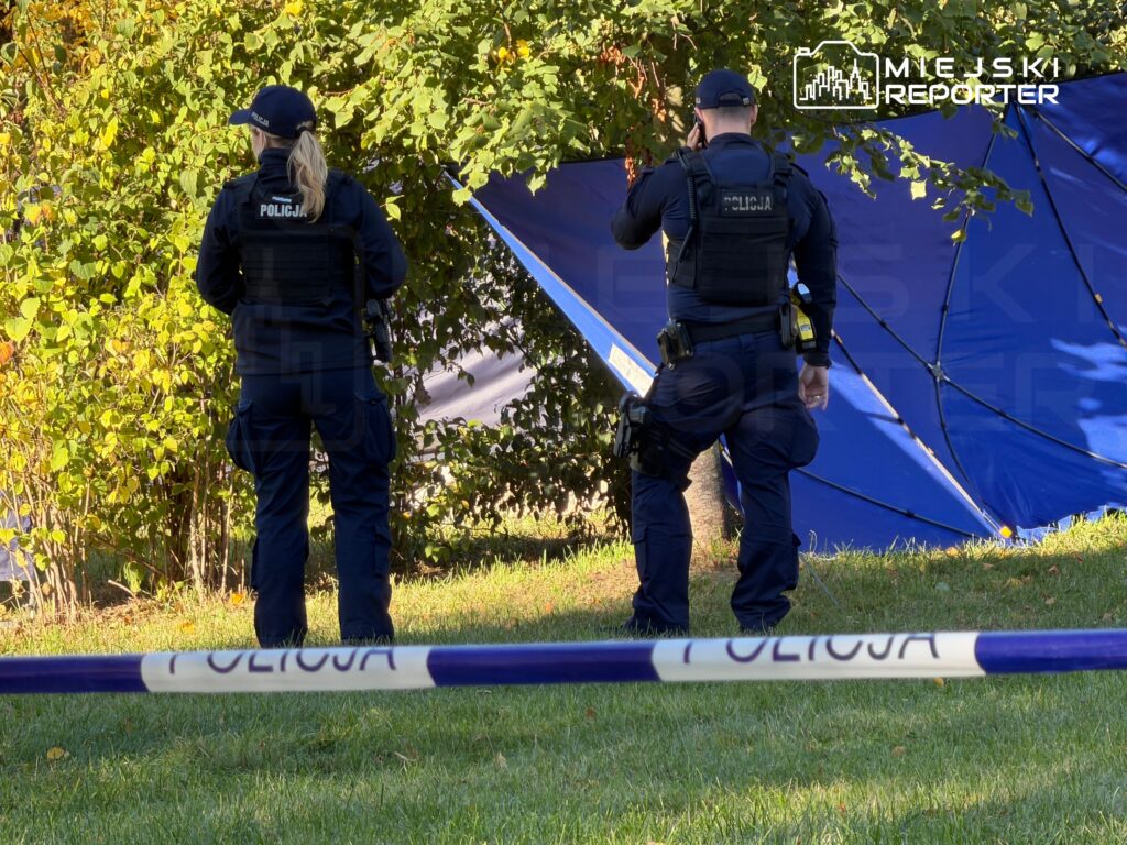 Dwoje policjantów stoi przy niebieskim namiocie za taśmą zabezpieczającą teren, wśród drzew.