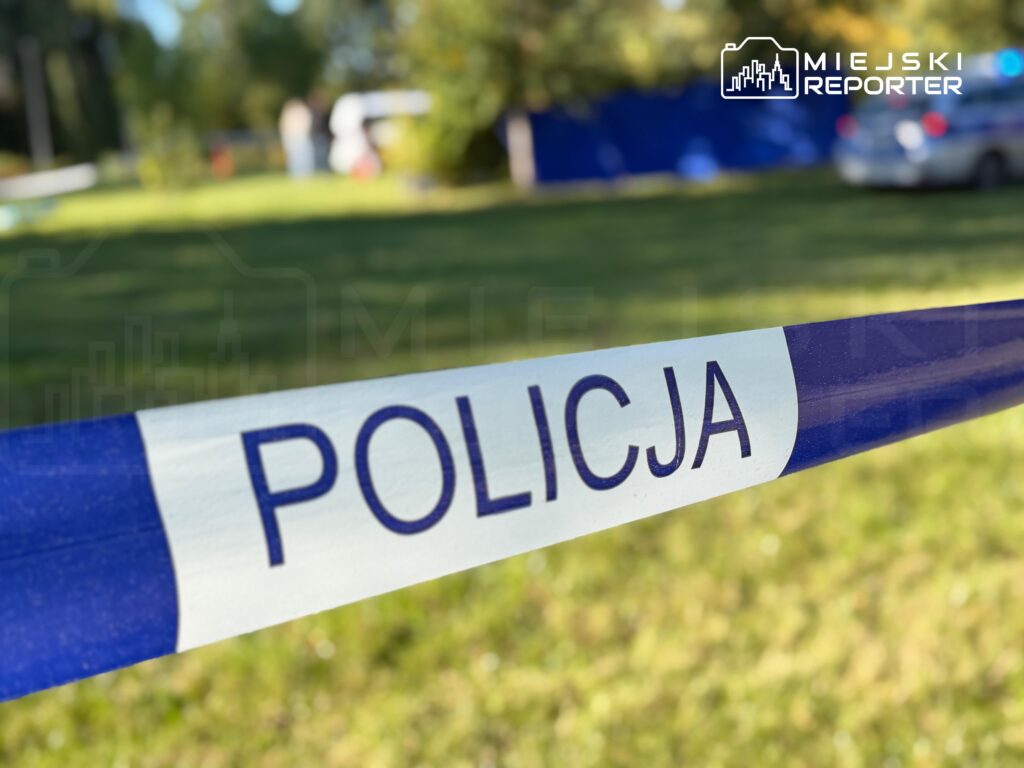 Taśma policyjna z napisem „POLICJA” odgradza trawnik w parku.