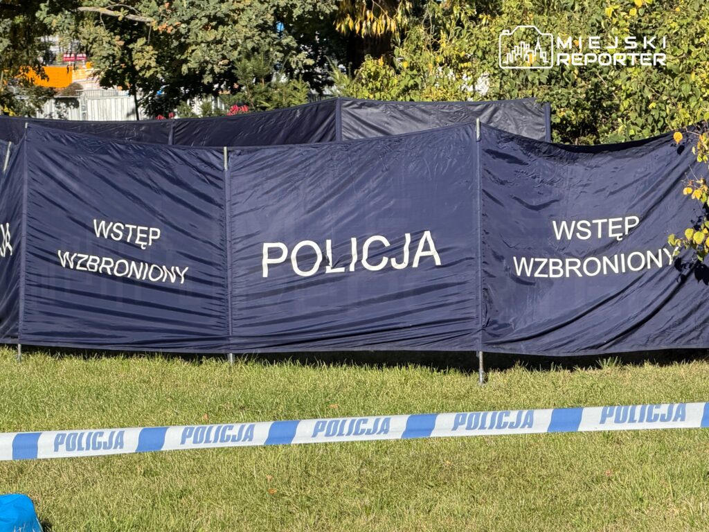 Policyjny parawan z napisem „POLICJA” i „WSTĘP WZBRONIONY” na trawie, za taśmą policyjną.