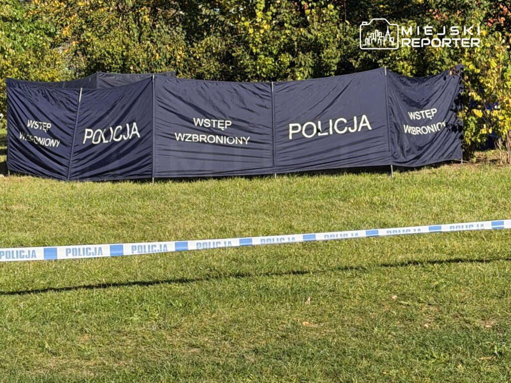 Parawan z napisem „POLICJA” i „WSTĘP WZBRONIONY” za policyjną taśmą na trawniku przy zaroślach.