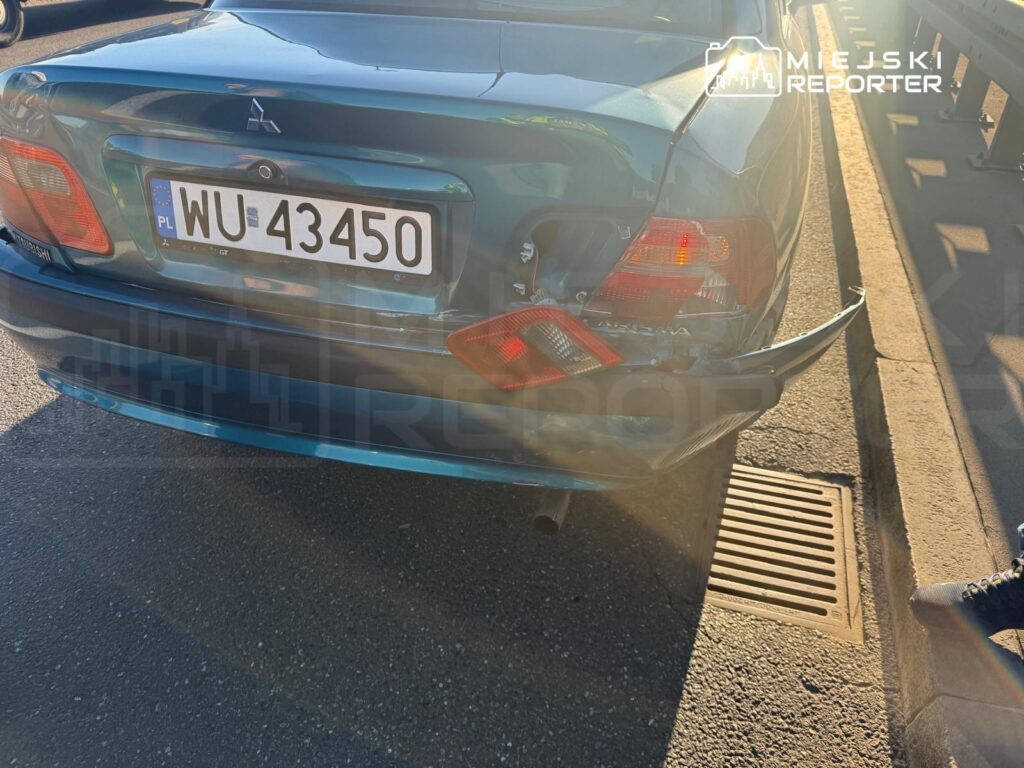 Tył niebieskiego Mitsubishi z uszkodzonym zderzakiem stoi przy krawężniku i kratce ściekowej.