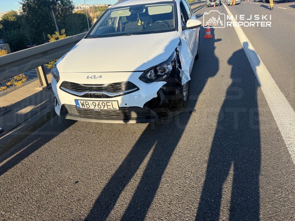 Uszkodzony biały samochód Kia stoi na pasie drogi przy barierze, a w oddali pracują służby.