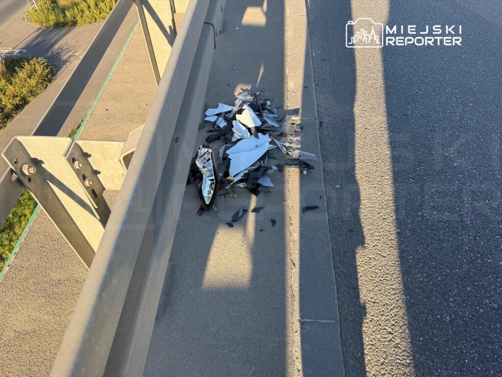 Rozbite części skutera leżą na chodniku przy metalowej barierze drogowej.