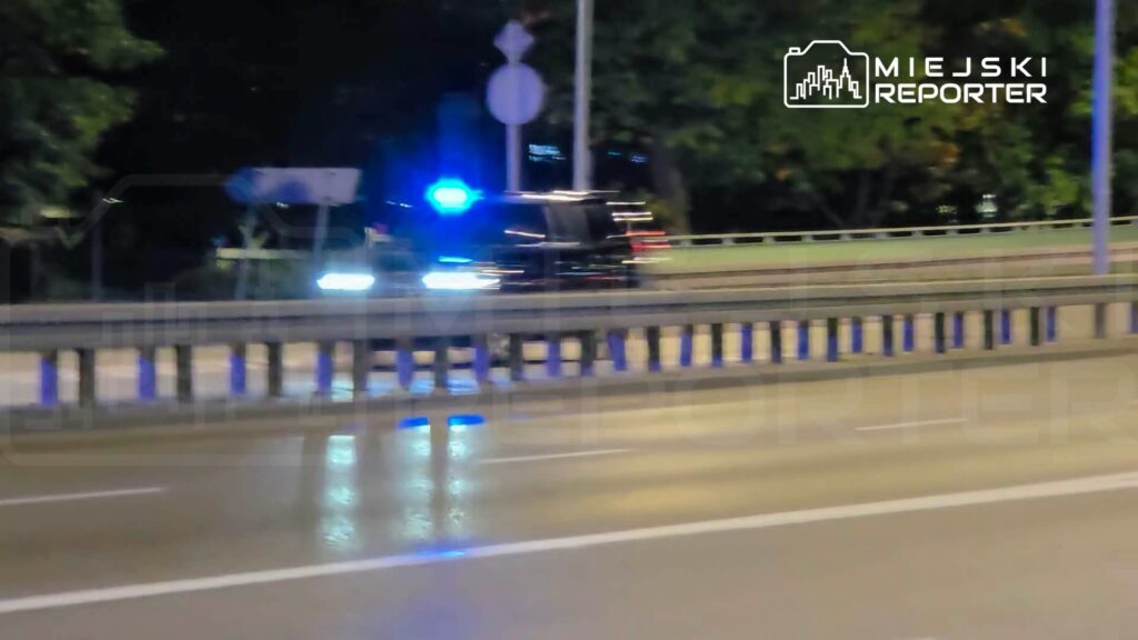 Samochód z włączonymi niebieskimi światłami jedzie nocą za barierą na mokrej drodze.