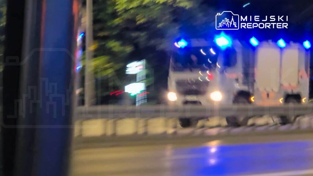 Ciężarówka z włączonymi niebieskimi światłami stoi nocą na jezdni za barierą drogową.