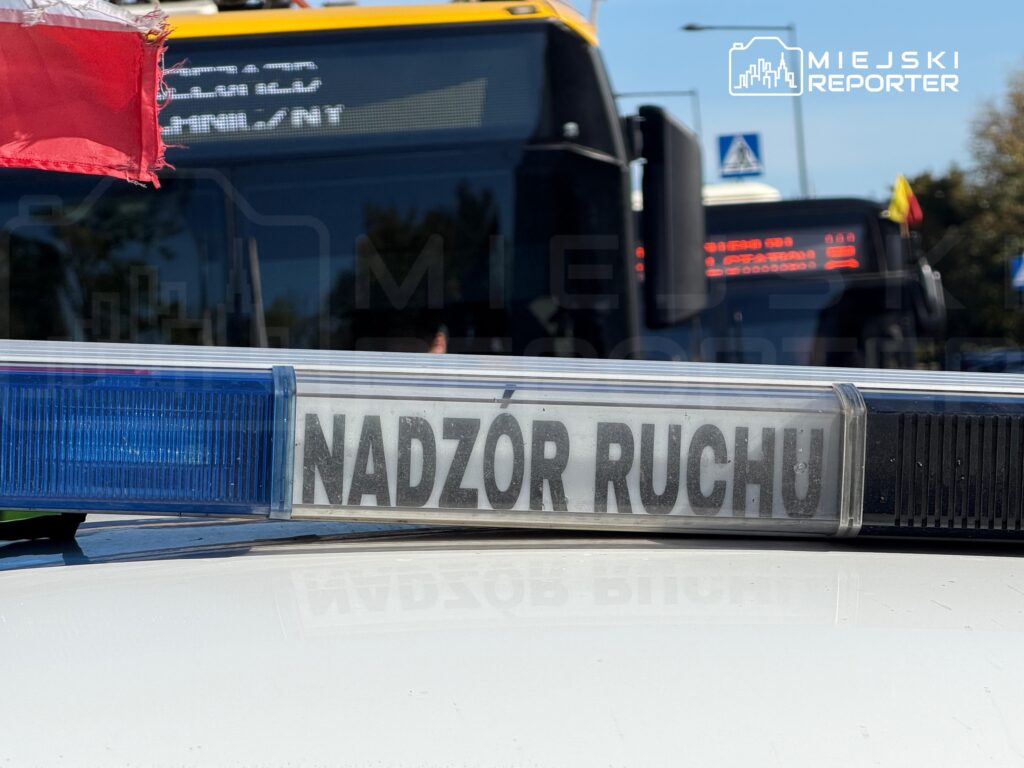 Belka świetlna na dachu radiowozu z napisem „NADZÓR RUCHU”, w tle autobusy miejskie i flaga.