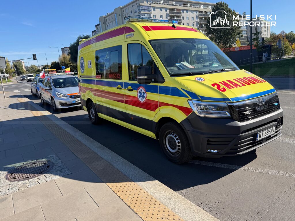 Żółty ambulans z napisem „AMBULANS” stoi przy miejskiej ulicy, za nim samochód osobowy.