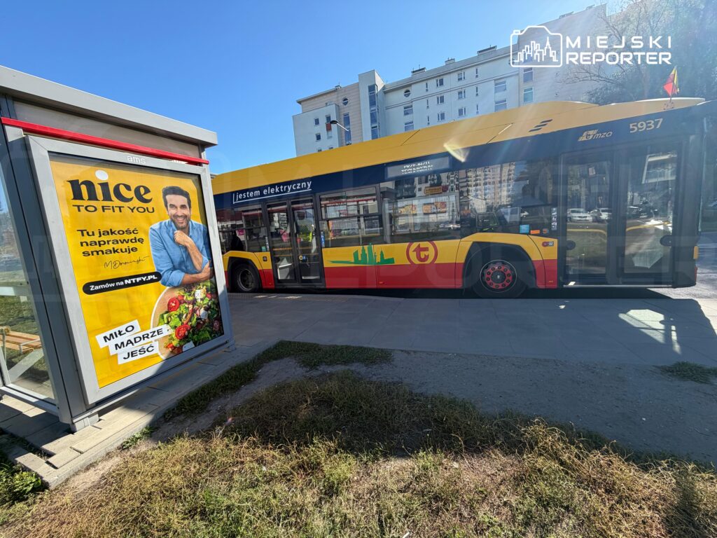 Żółto-czerwony autobus elektryczny stoi przy przystanku; na boku widnieje napis „Jestem elektryczny”.