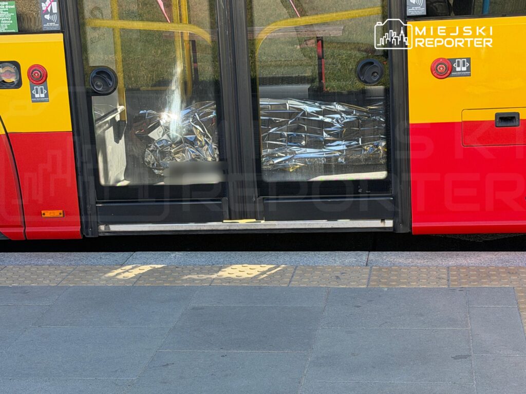 Uszkodzone drzwi tramwaju z wybitymi szybami, stojącego przy peronie przystankowym.