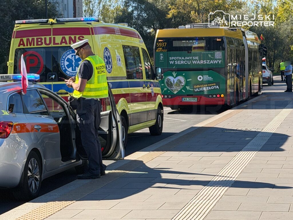 Ratownik w odblaskowej kamizelce stoi przy otwartych drzwiach samochodu obok karetki i autobusu na przystanku.