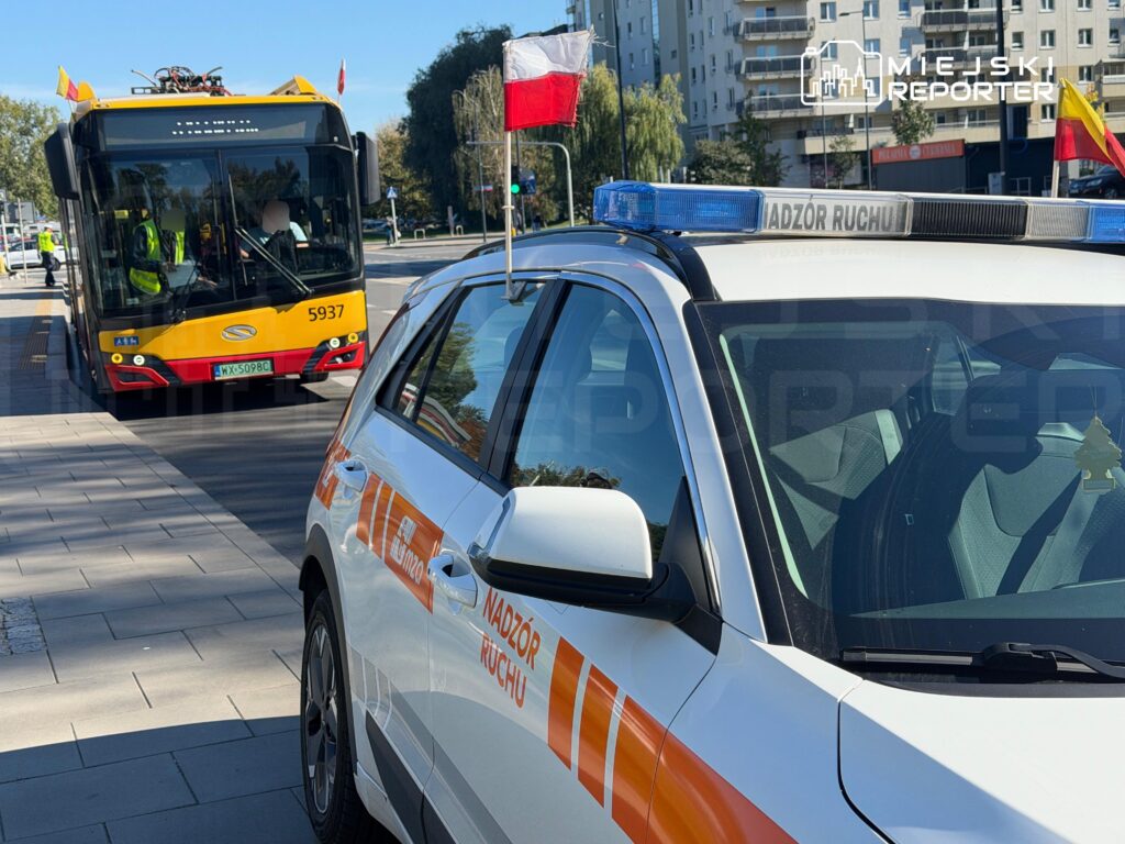 Oznakowany samochód nadzoru ruchu z biało-czerwoną flagą stoi przy chodniku, obok nadjeżdża żółto-czerwony autobus.