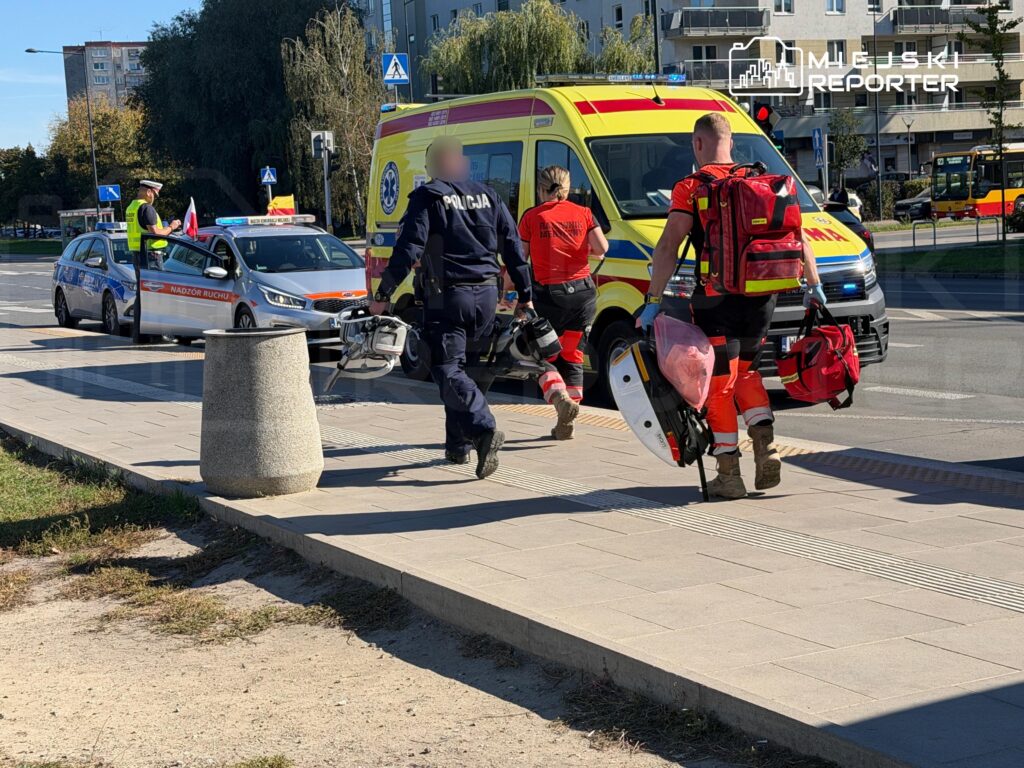 Policjant i ratownicy medyczni niosą sprzęt obok karetki na miejskiej ulicy, przy radiowozie i skrzyżowaniu.