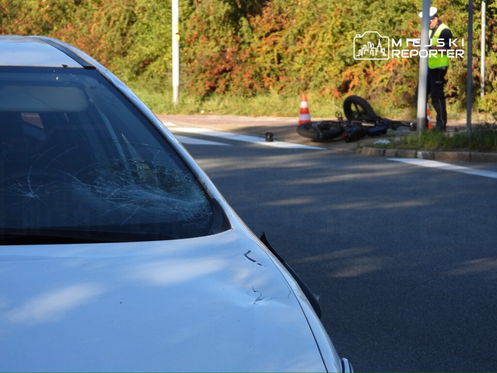 Uszkodzony samochód z pękniętą szybą stoi na jezdni obok przewróconego motocykla, pachołka i funkcjonariusza drogówki.