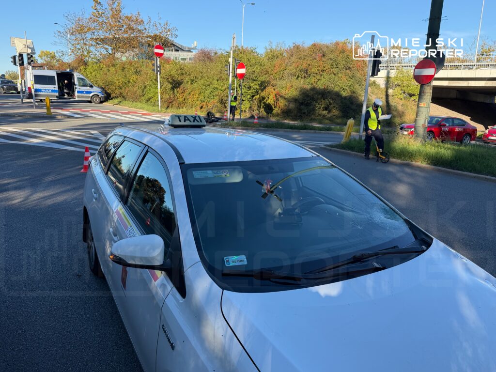 Biała taksówka stoi przy skrzyżowaniu, a policjant w kamizelce odblaskowej zabezpiecza miejsce zdarzenia.