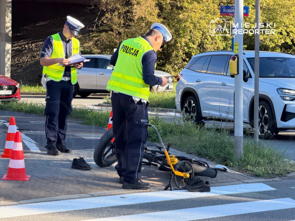 Policjanci w odblaskowych kamizelkach oglądają przewrócony rower przy przejściu dla pieszych, obok stoją pachołki drogowe.